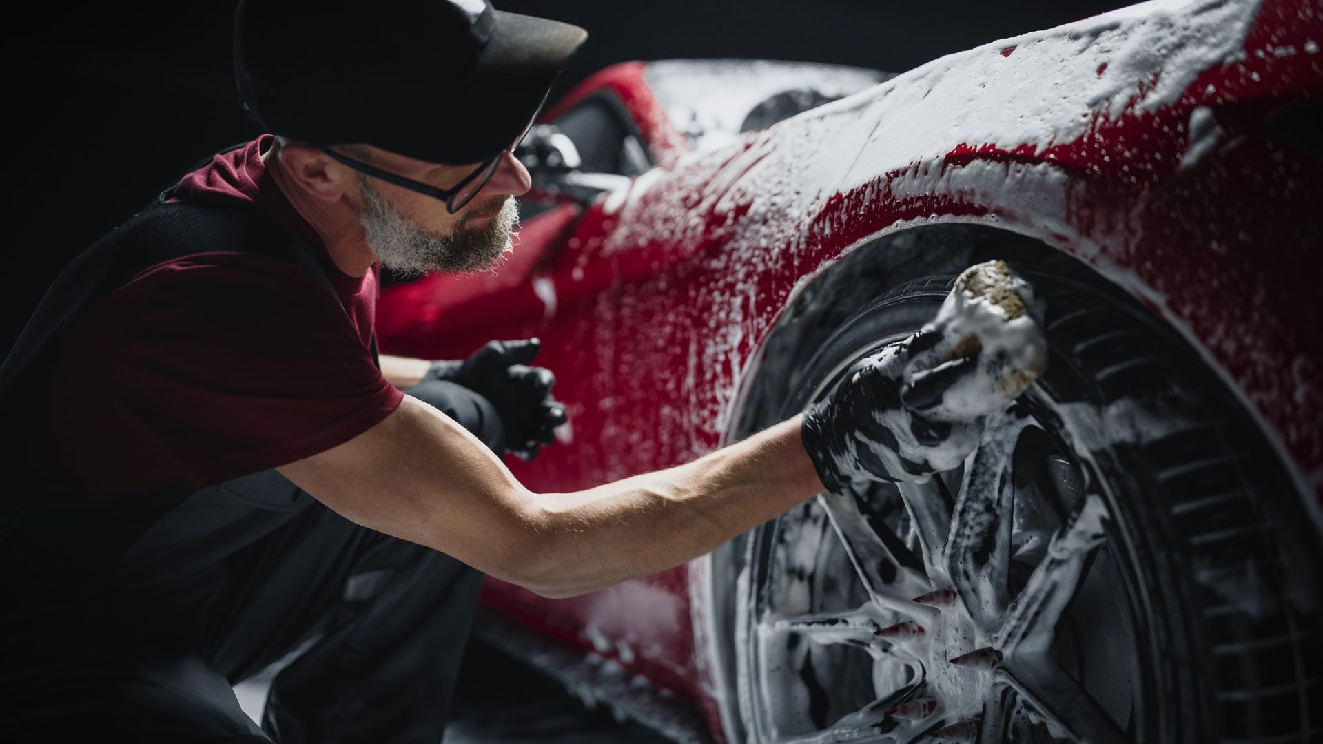 Man washes a red car's tire with a soapy sponge. Dark background.