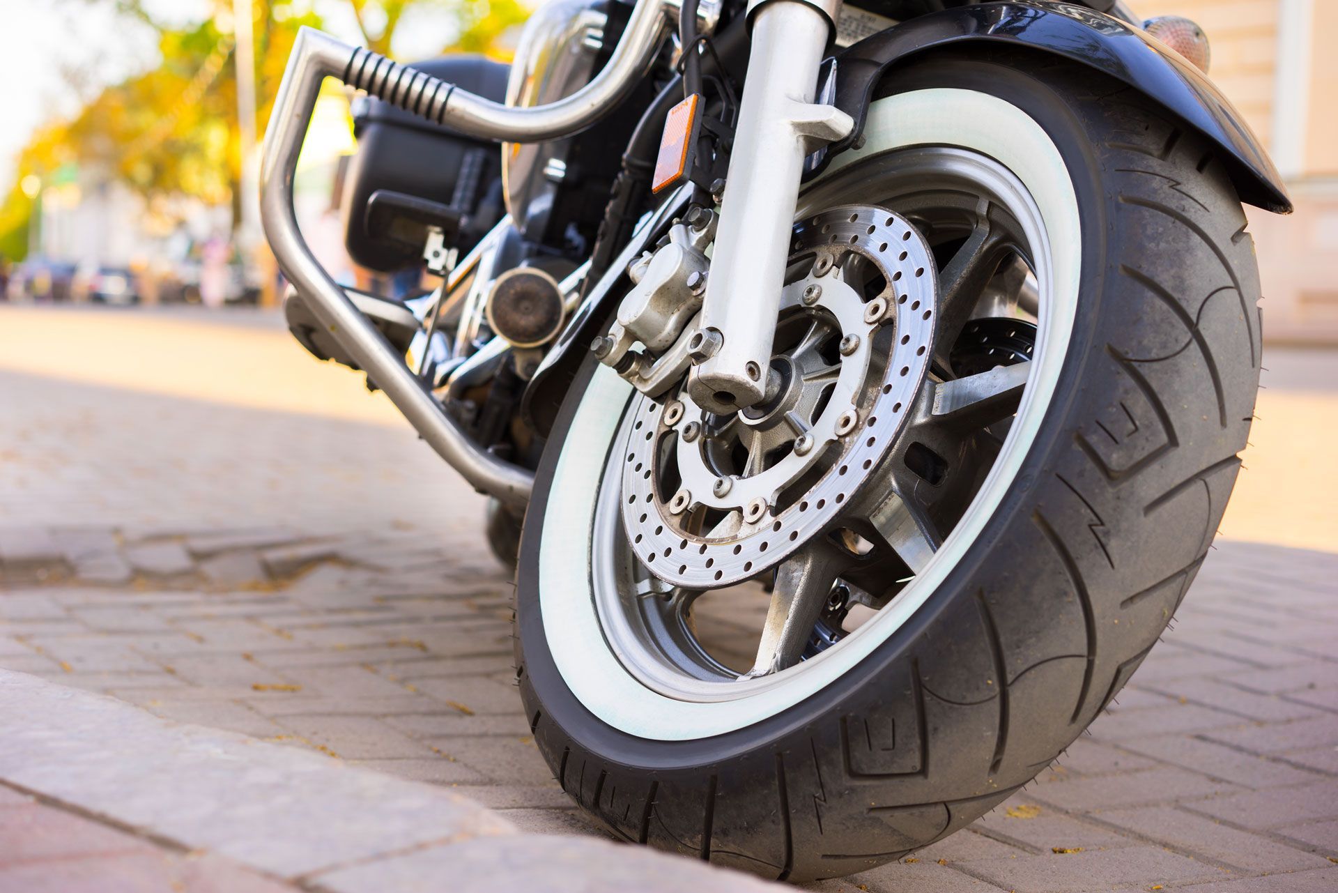Close-up of a black motorcycle front wheel with white sidewall tire parked on a brick-paved street.