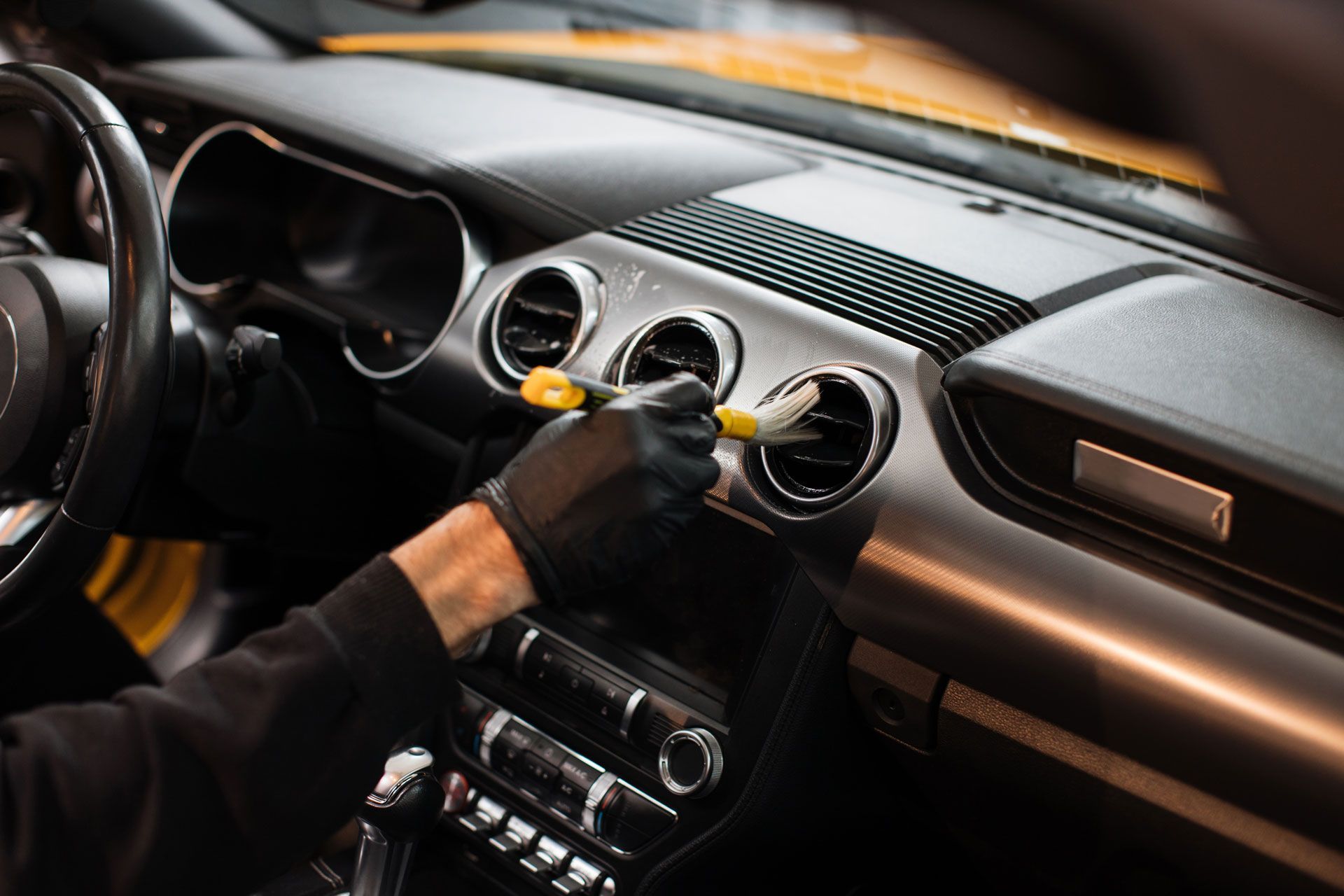 Black-gloved hand cleaning a car's dashboard vents with a brush. Interior of a car is visible.