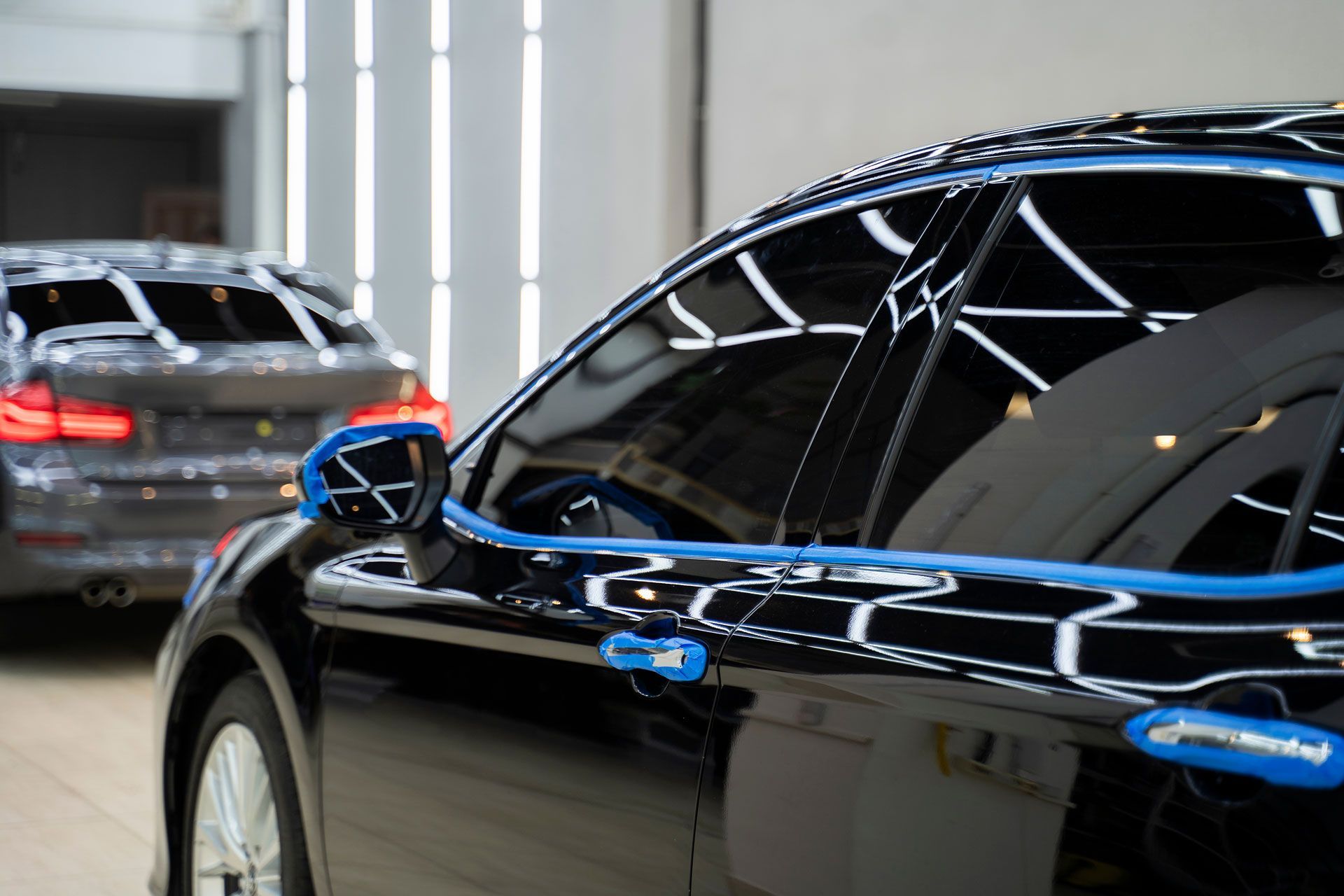 Black car being detailed in a garage; blue tape around windows, another car blurred in background.