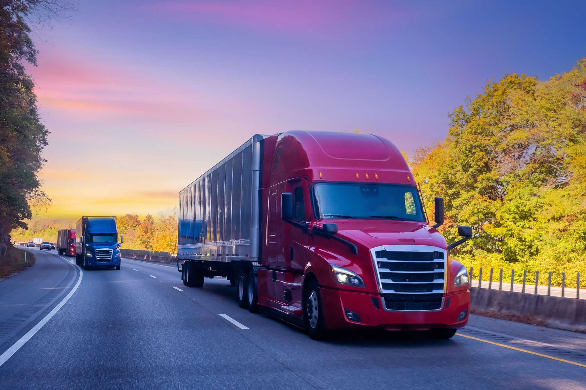 Red semi-truck on highway, blue truck in background, under sunset sky, trees along the roadside.