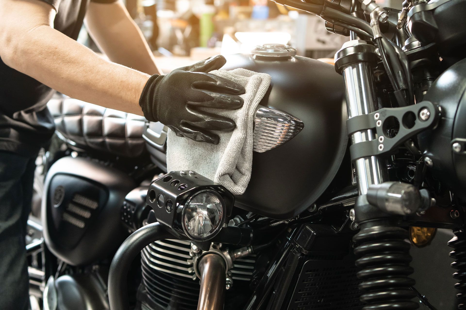 A person in gloves wipes a black motorcycle's gas tank with a cloth inside a garage.