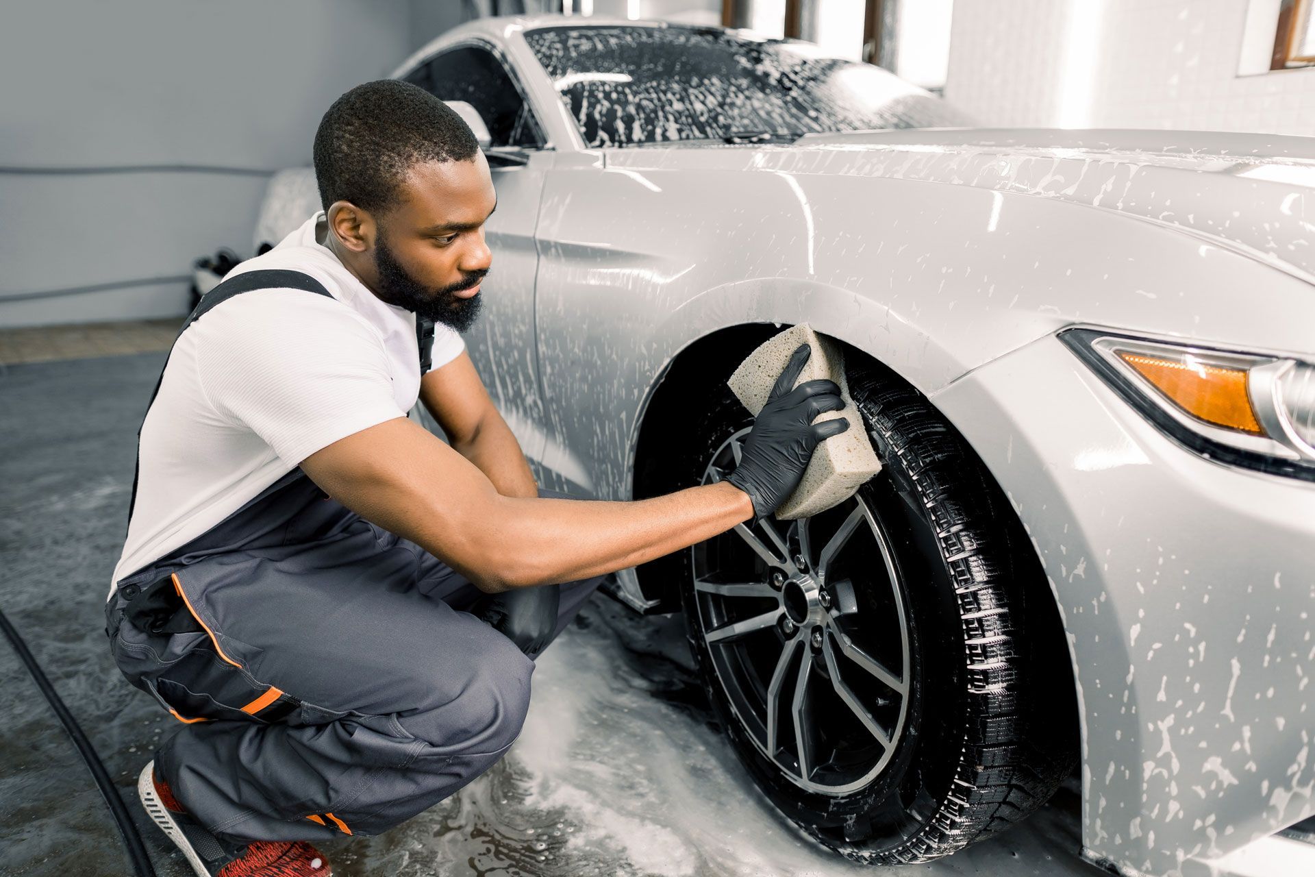 Person washing a silver car tire with a sponge, in a car wash bay.