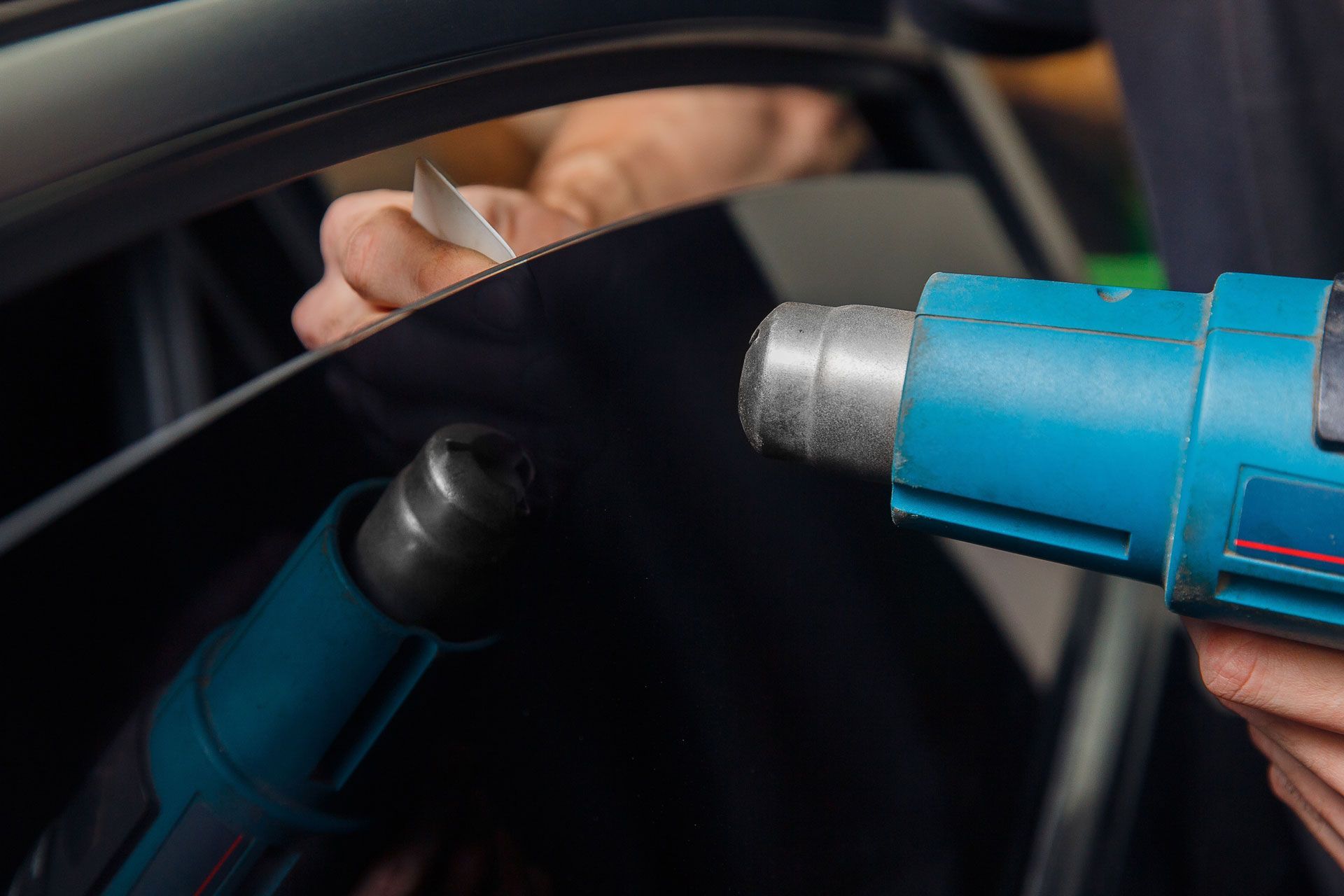 Person applying tint to a car window with a heat gun, close-up. Blue heat gun, black window tint.