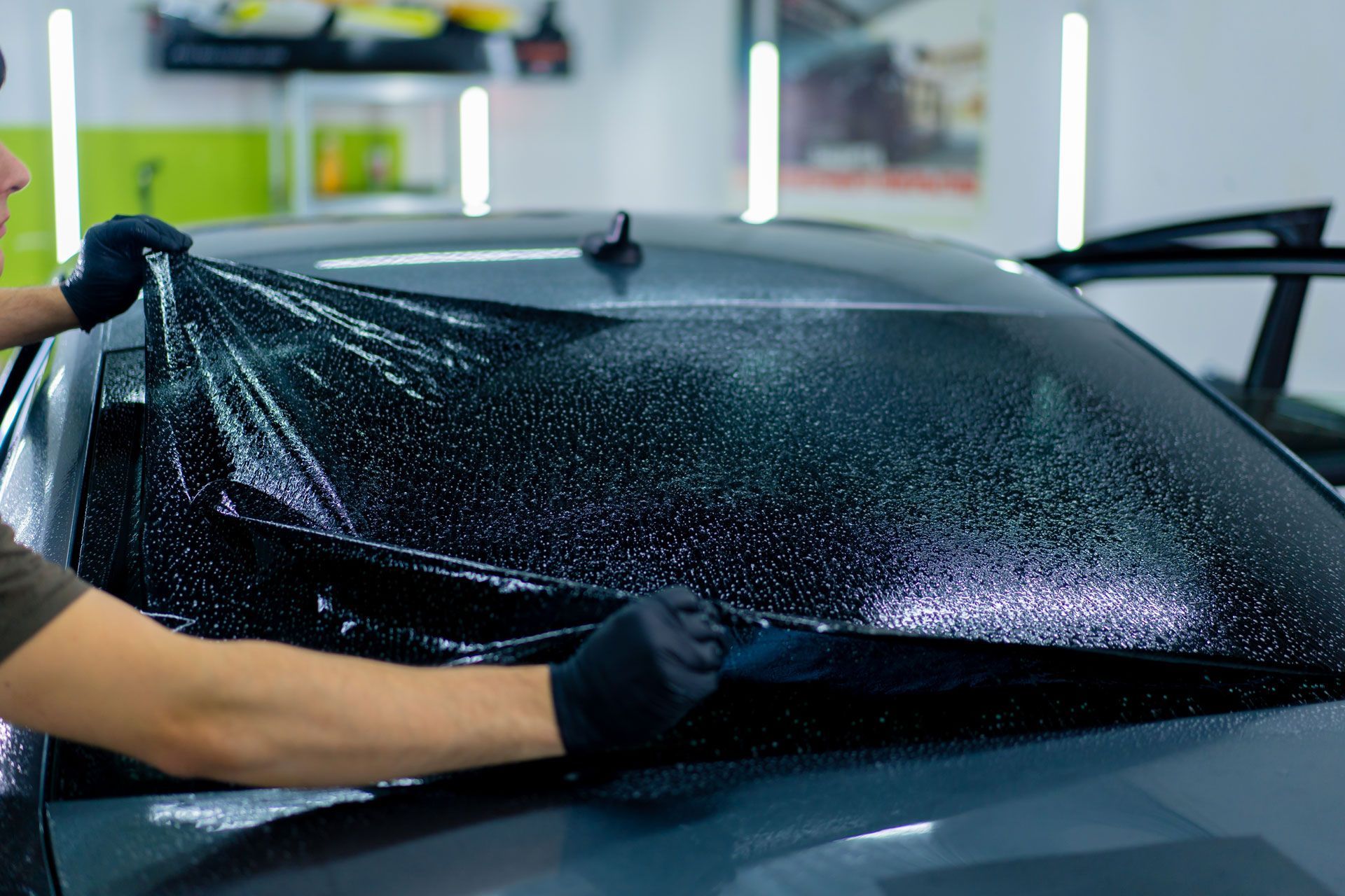 Person applying tinted film to a car's rear window indoors. Black gloves, wet window, gray car.