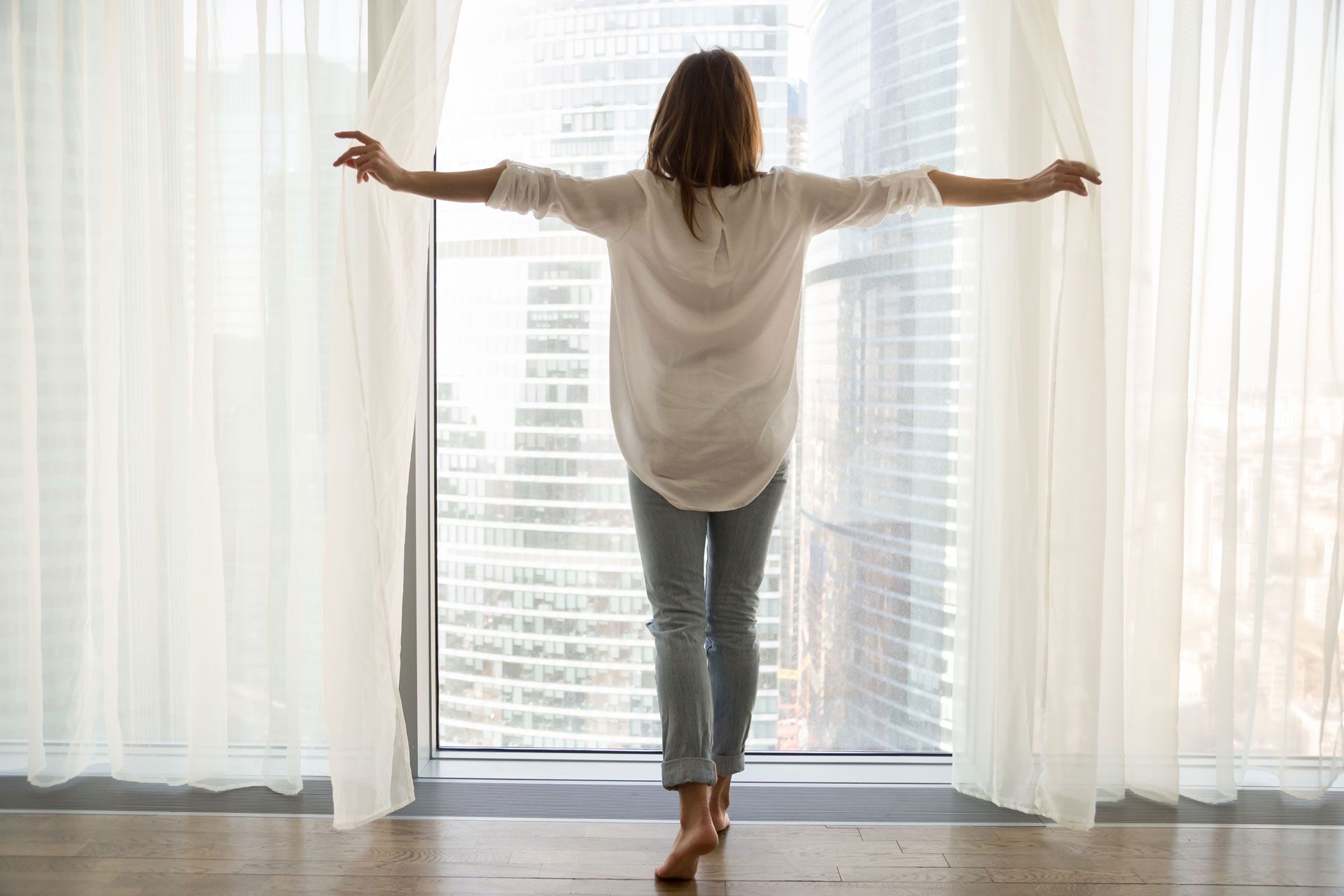 A person stands barefoot in a bright room, arms outstretched while opening sheer white curtains to reveal a city view.