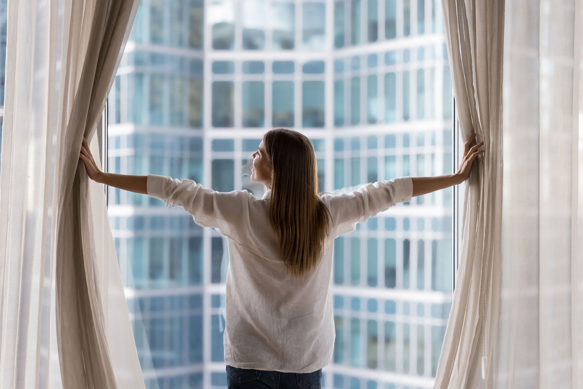 A person stands facing a window, arms outstretched, pulling back sheer white curtains to reveal a large office building.