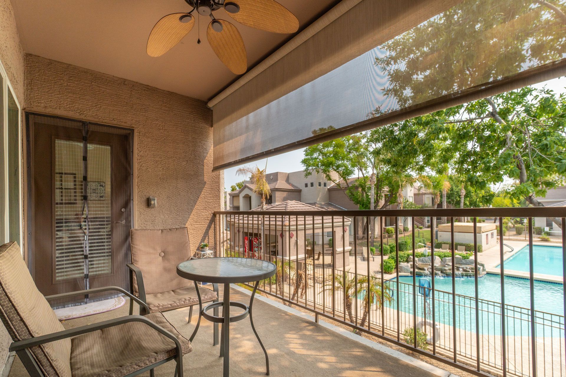 A balcony with a patio chair, round table, and ceiling fan overlooking a swimming pool and residential buildings.