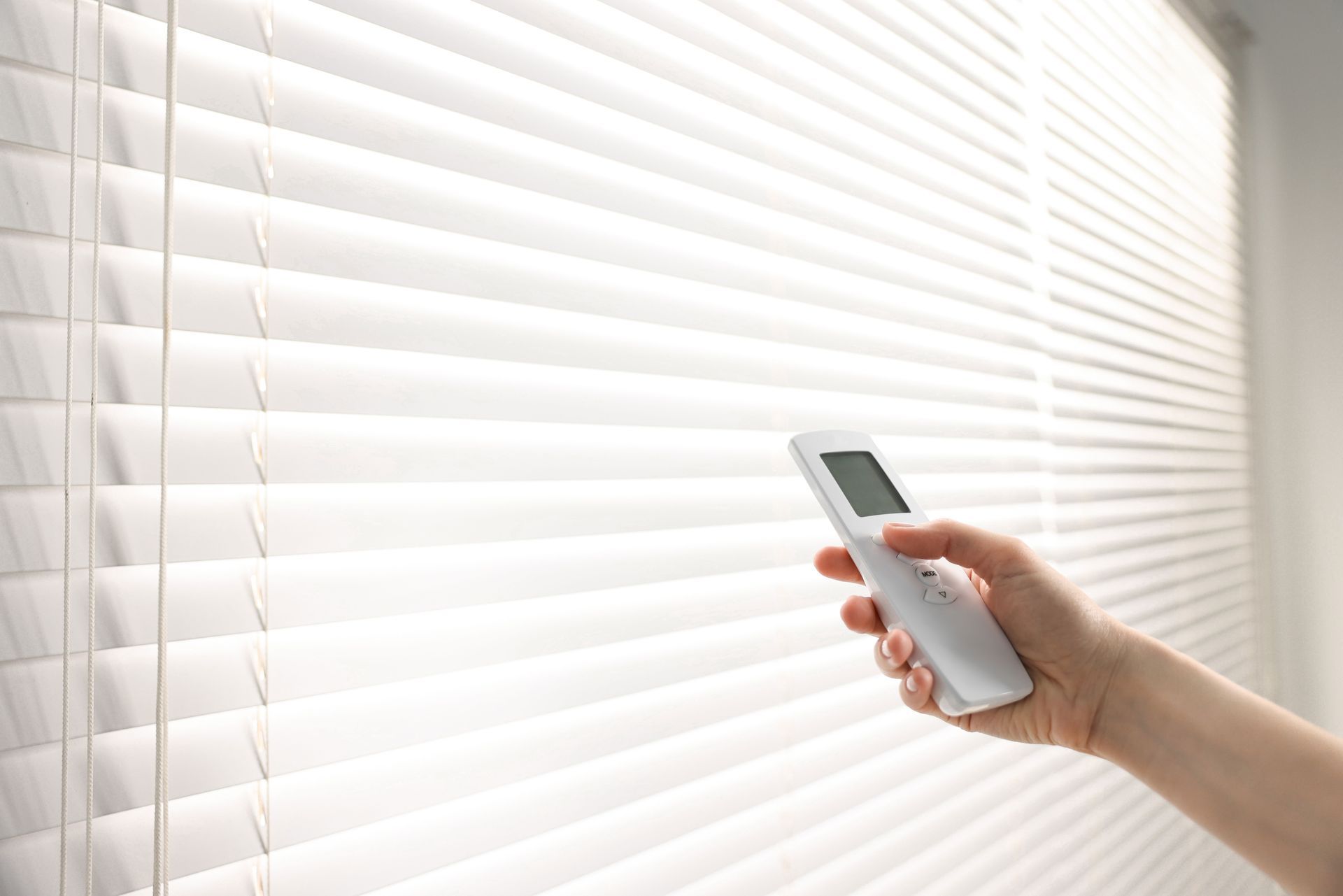 Hand holding a remote control adjusting white, horizontal blinds on a window.