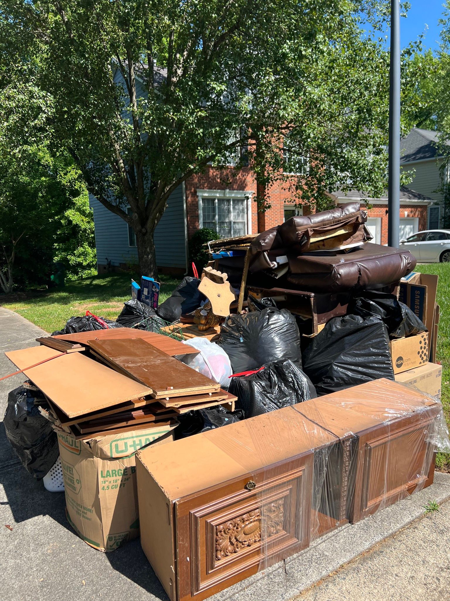A large pile of assorted trash, cardboard, and discarded furniture waiting on a curb in front of a brick house.