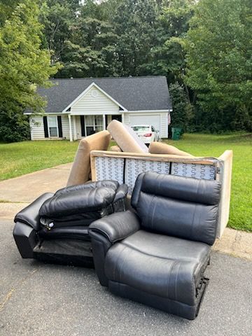 Discarded black leather couch cushions and armchair pieces on a driveway in front of a suburban house.