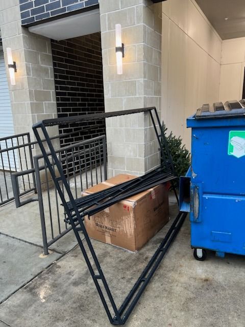 A black metal A-frame display rack stands outdoors on a concrete sidewalk beside a large blue dumpster and cardboard box.