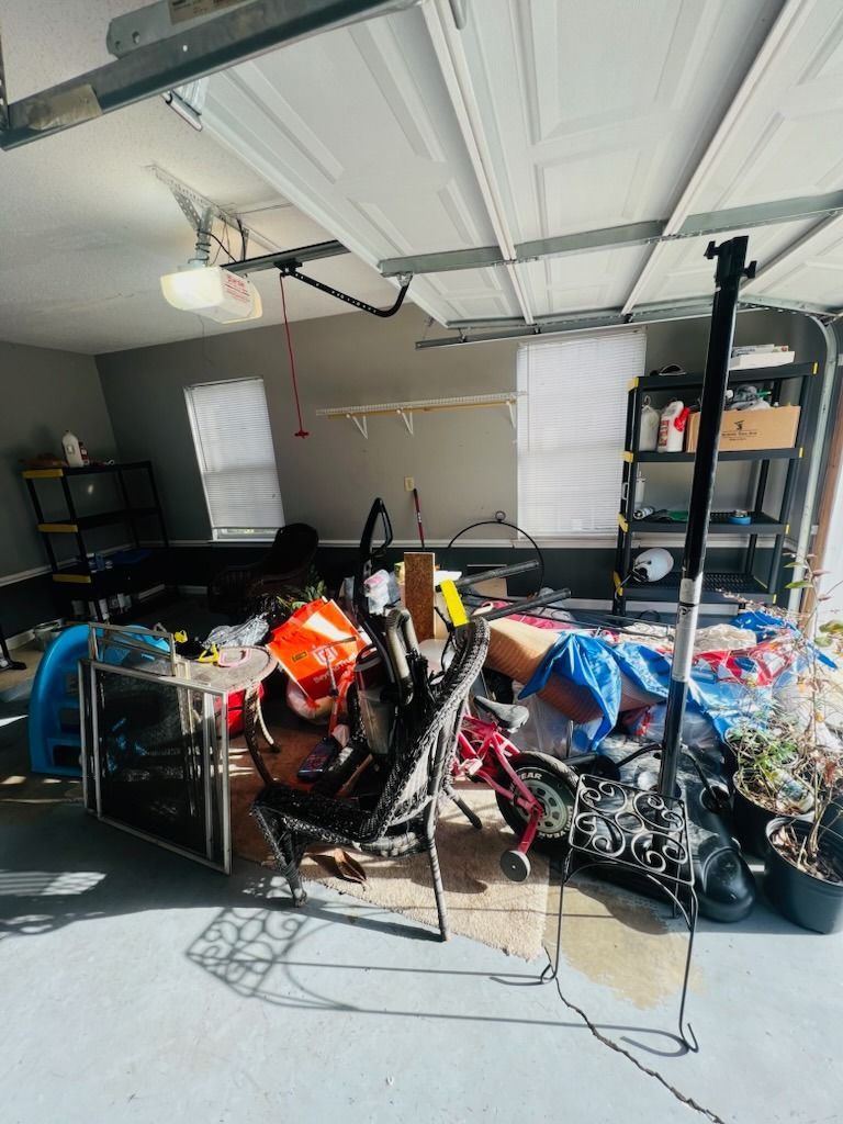 A cluttered garage with a metal shelving unit, a desk, various stored items, a tripod, and potted plants.