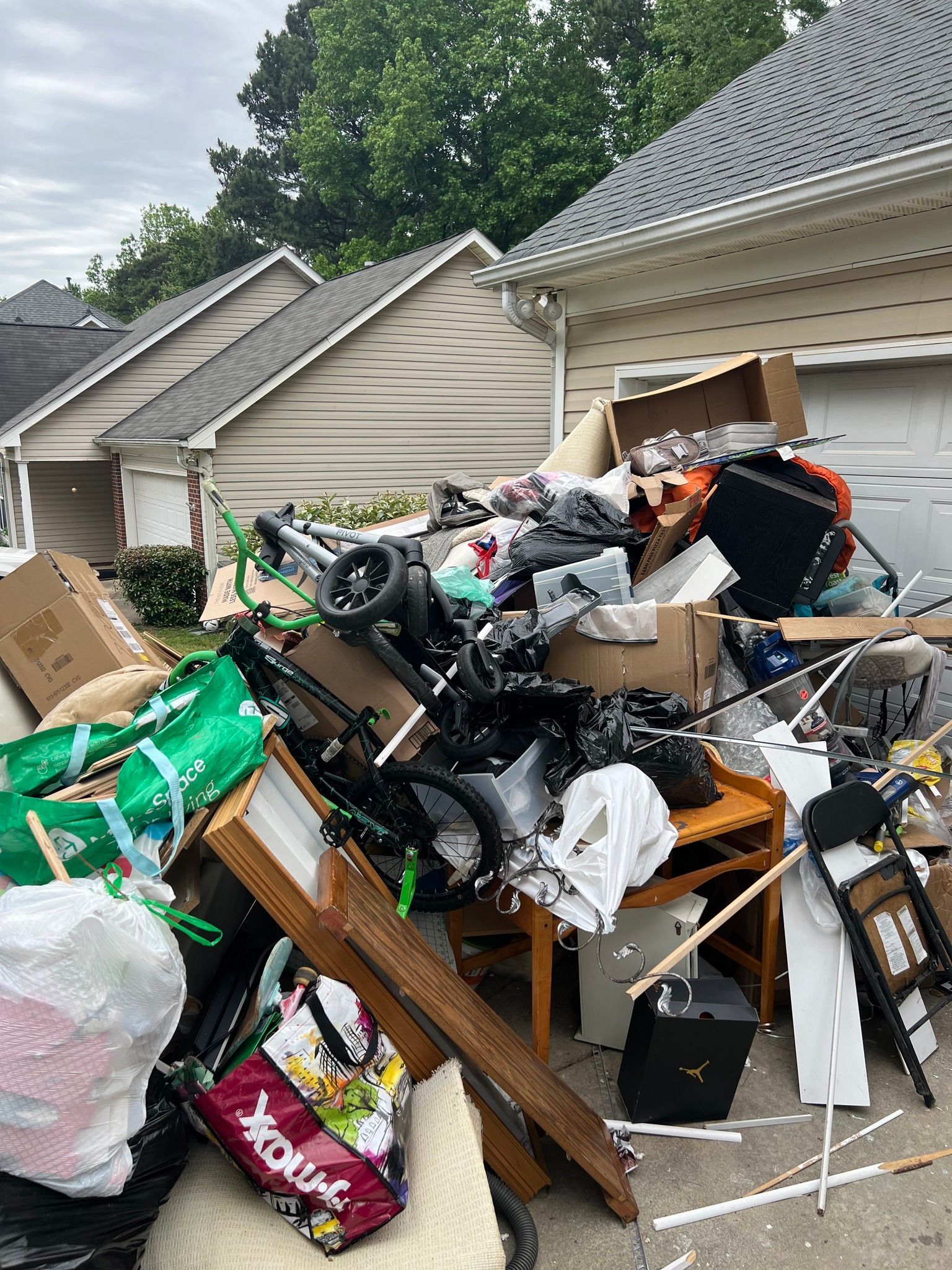 A large pile of household junk and debris sits on a suburban driveway in front of a garage.