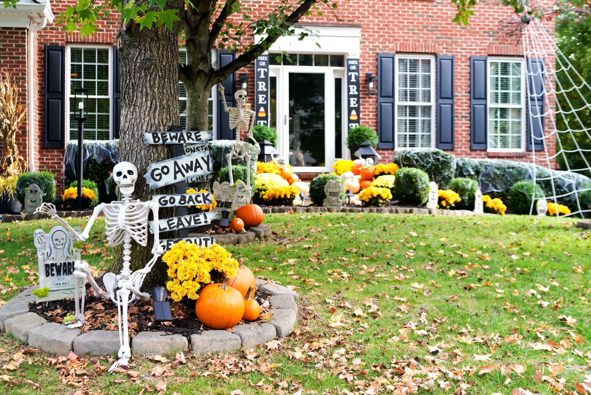 A brick home with Halloween lawn decorations, including skeletons, pumpkins, fall flowers, and a large spider web.