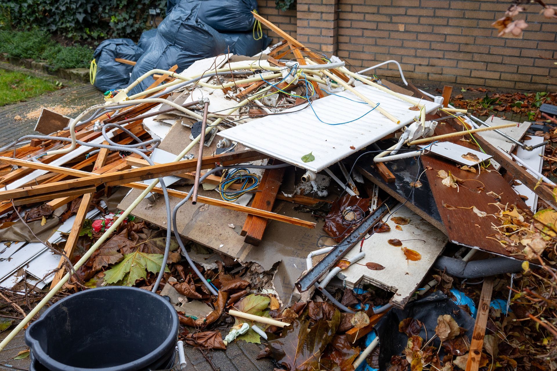 Pile of demolition debris with broken wood, drywall, and a blue bucket in a yard near a brick wall