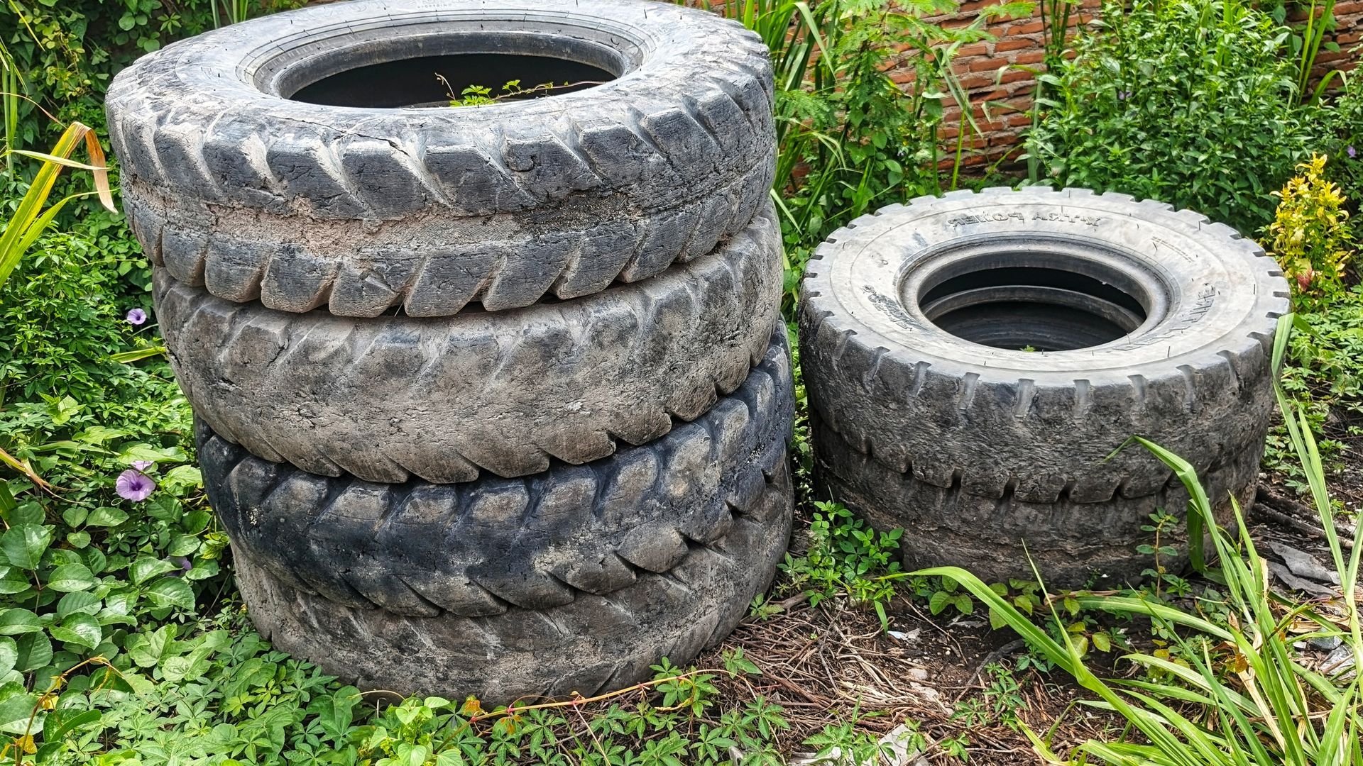 A stack of three weathered, dark-colored tires sits next to a single tire in a grassy garden area.
