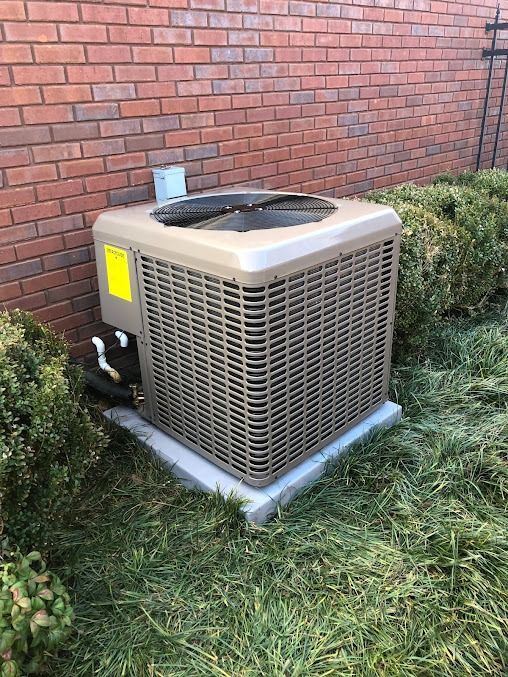 Air conditioning unit on a concrete pad near a brick wall and green shrubs.