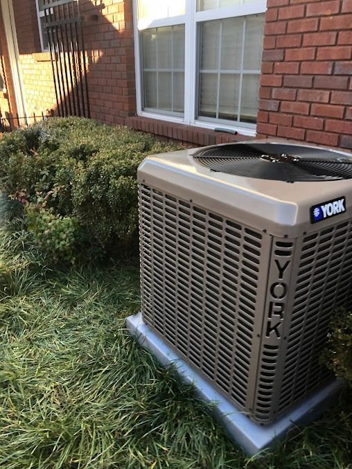 York air conditioner unit outside a brick building, nestled in a grassy area with greenery and a window visible.
