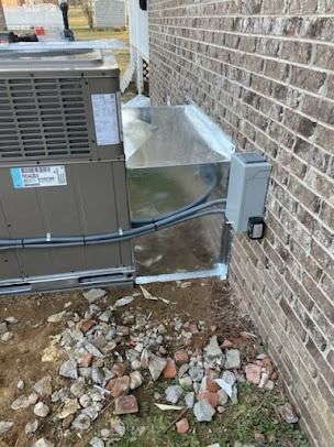 HVAC unit beside a brick wall. Galvanized metal ductwork connects unit to the wall. Power box attached. Ground littered with brick pieces.