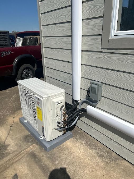 An air conditioning unit on a concrete pad outside a building, with white piping running along the wall.
