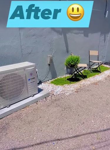 Air conditioner next to a small gravel and artificial grass patio with seating and a potted plant.