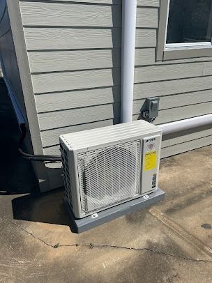 An air conditioning unit on a concrete pad near a building with gray siding and a white pipe.