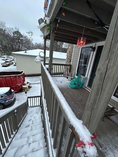 Snow-covered deck and stairs leading down to a snowy yard, with a white building and parked cars in the distance.
