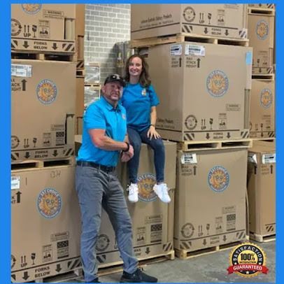 Two people pose on pallets of boxes in a warehouse. They wear matching blue shirts, and smile. Boxes are stacked high.