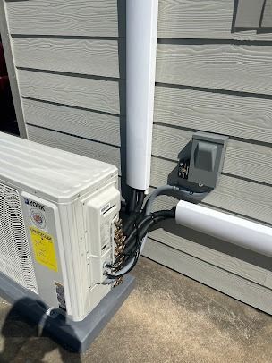 Outdoor AC unit with conduit and electrical box mounted on a gray-sided building.