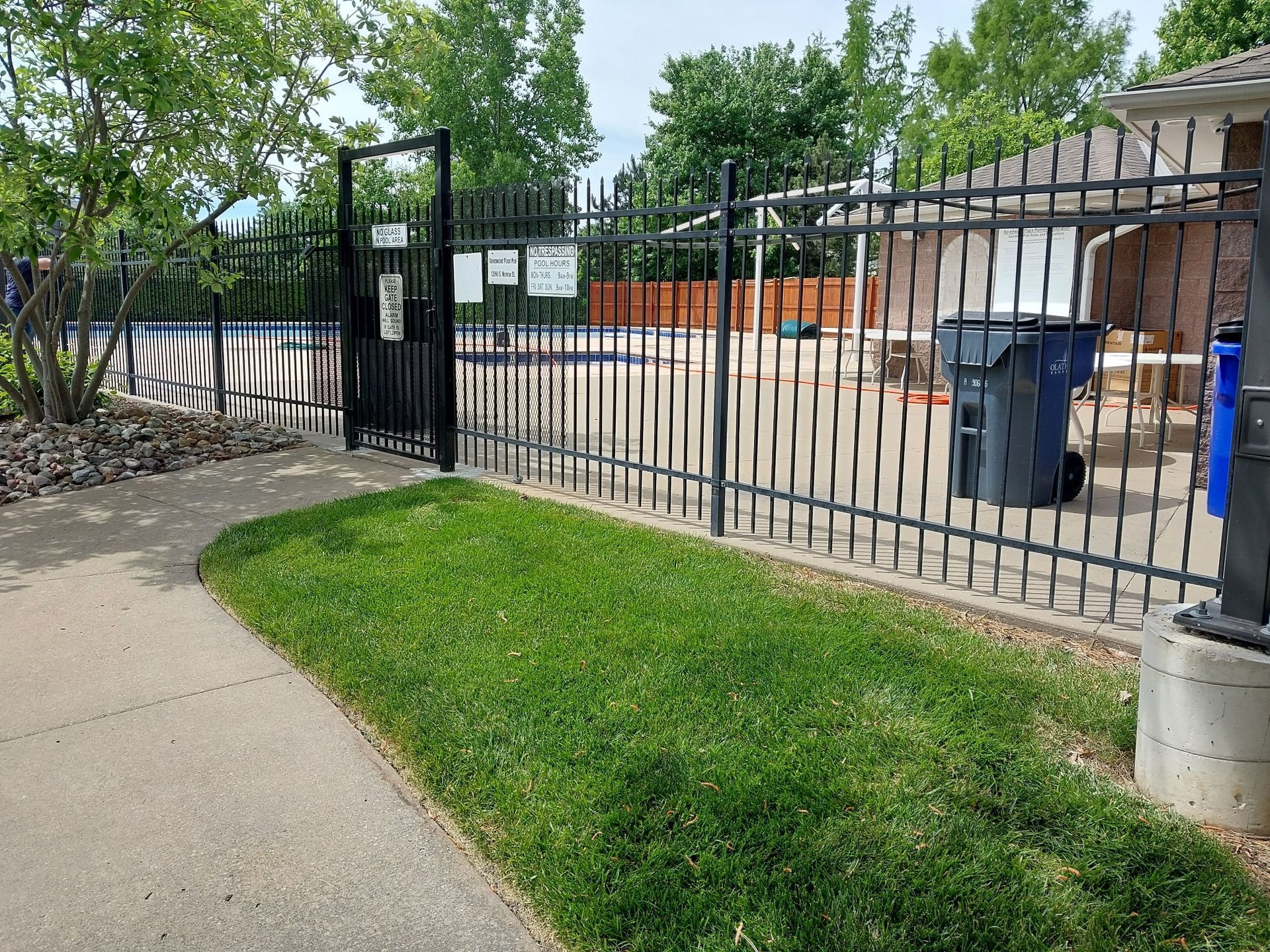 A metal fence surrounds a swimming pool with a trash can in front of it.