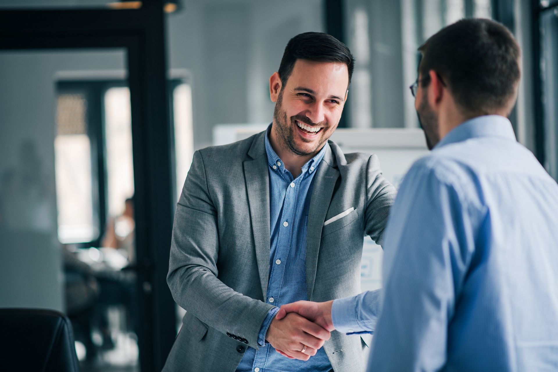 Two men shaking hands and smiling in an office, one in a grey suit, the other in a blue shirt.