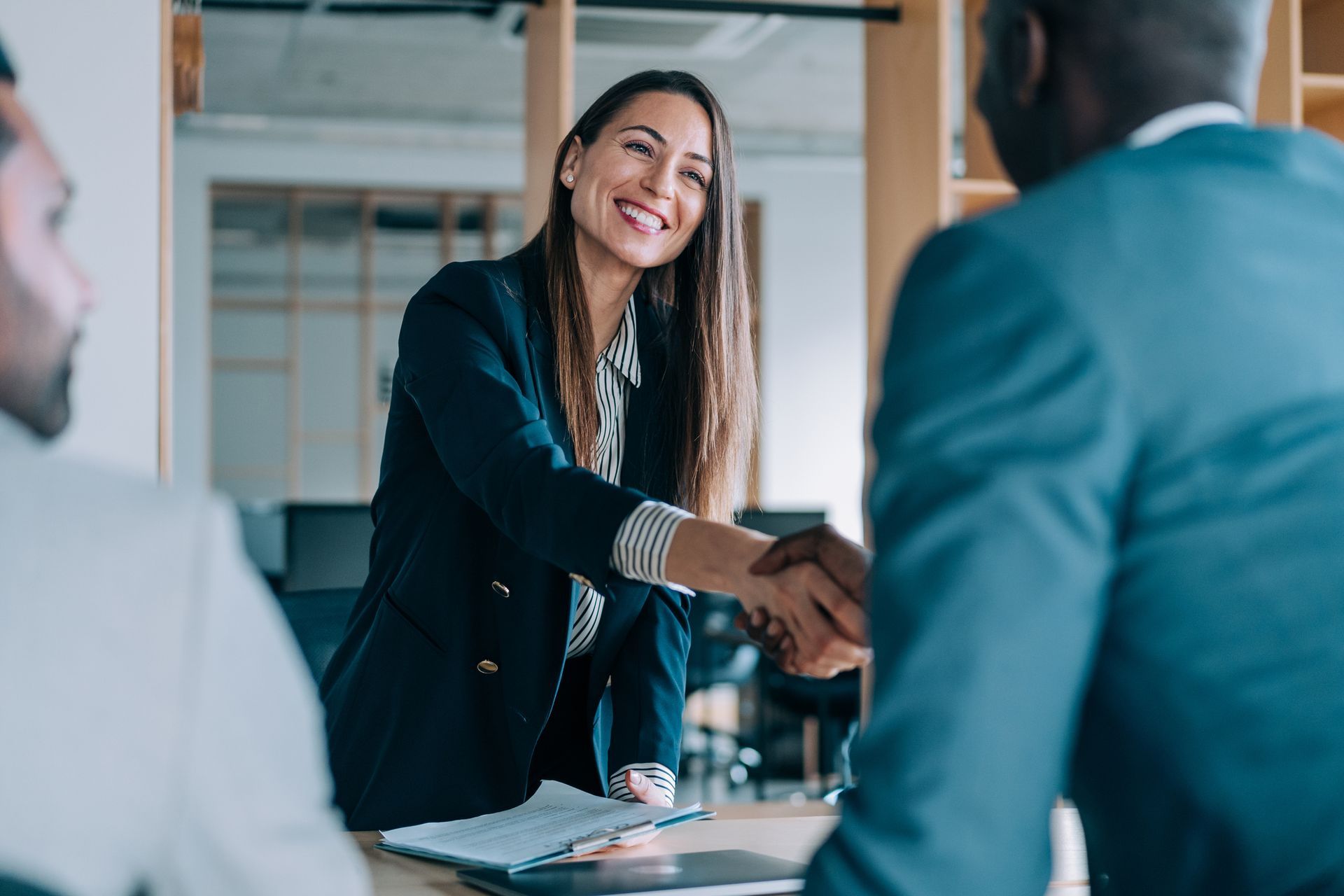Woman in blazer shakes hands with a man, smiling, in an office setting. Two other men are present.
