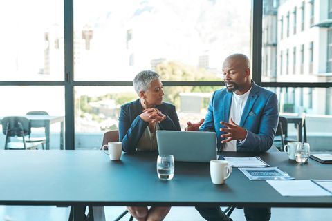 Two business people, a man and woman, at a table with a laptop and papers, discussing.