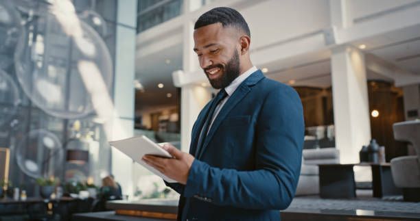 Man in blue suit smiles while using a tablet in a modern, bright building lobby.
