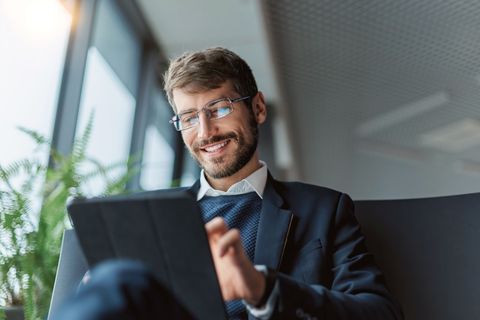 Man in glasses smiles, using a tablet. He wears a suit jacket and sweater, seated by a window.