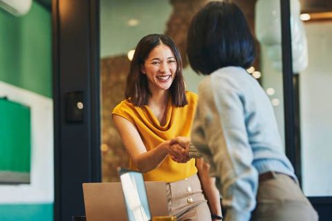 Two women shaking hands, smiling, in an office setting. One wears yellow, the other light blue.