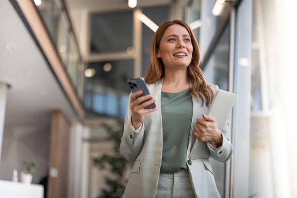 Woman in a light suit walks in an office, holding a phone and folder, smiling.