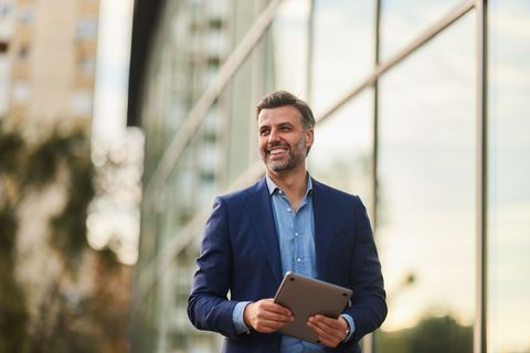 Man in suit smiling, holding tablet, standing near glass building.
