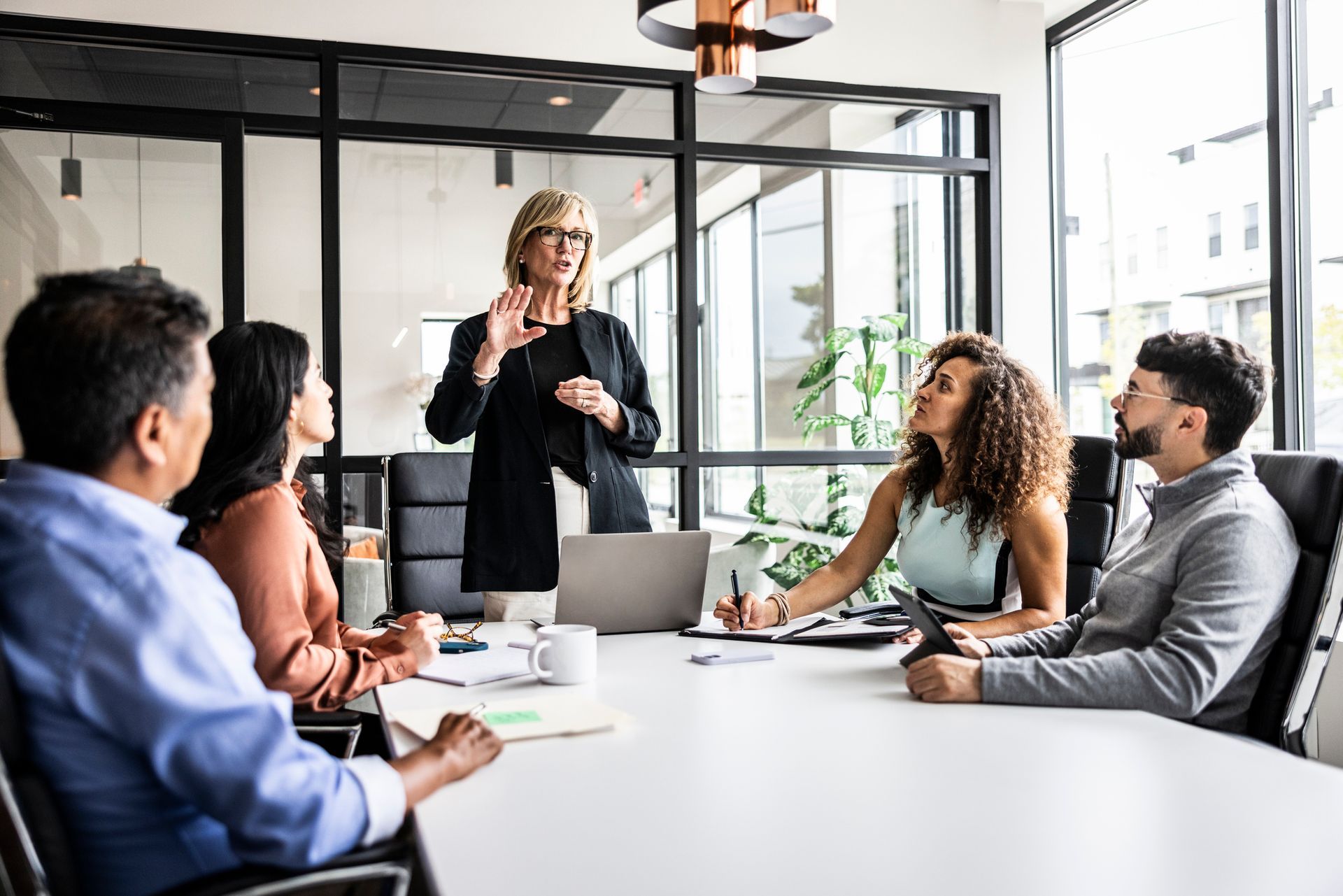 A woman in a suit leads a meeting at a table; others look on and listen in a bright office.