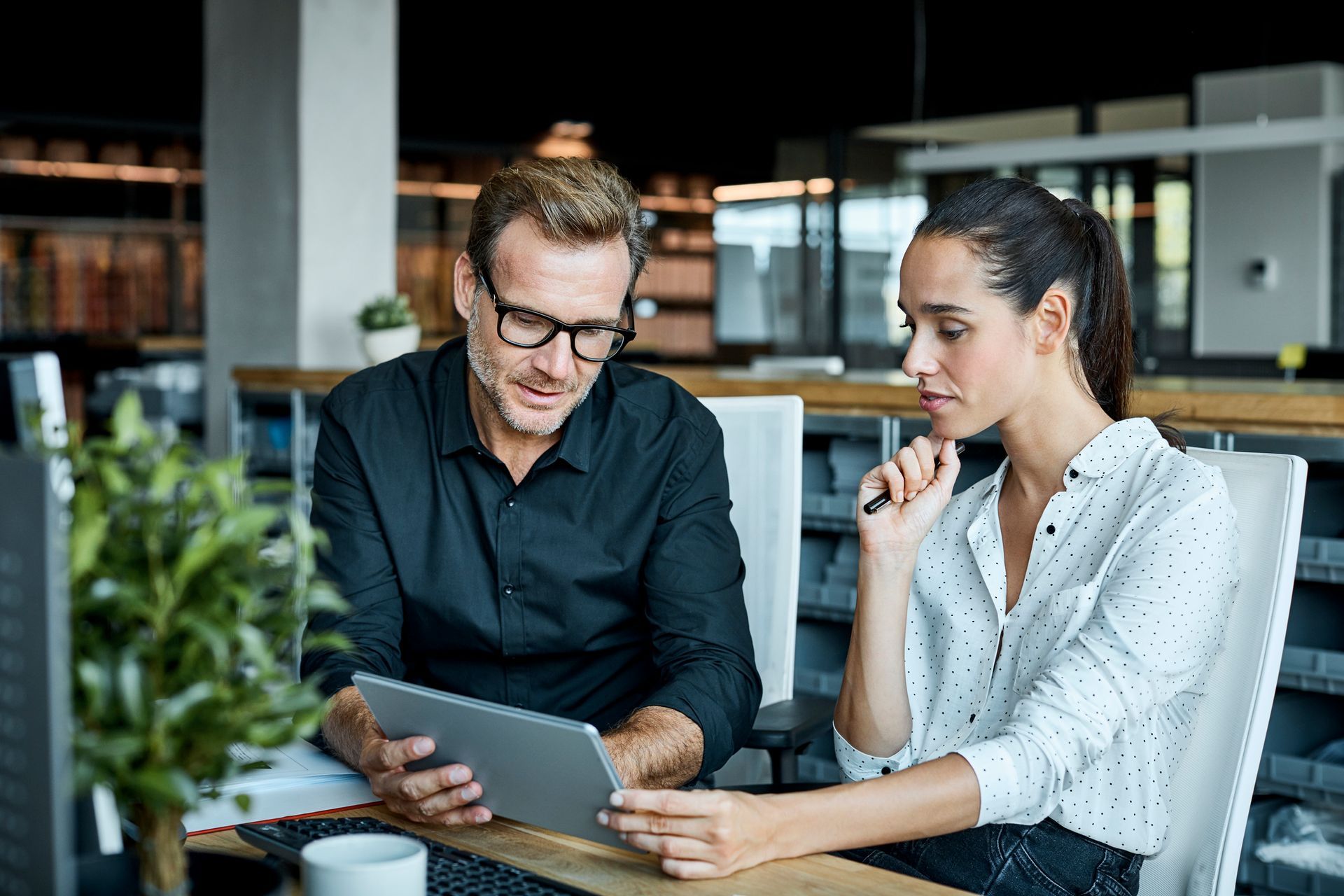 Man and woman looking at tablet in an office setting. Man points to screen, woman holds pen, both focused.
