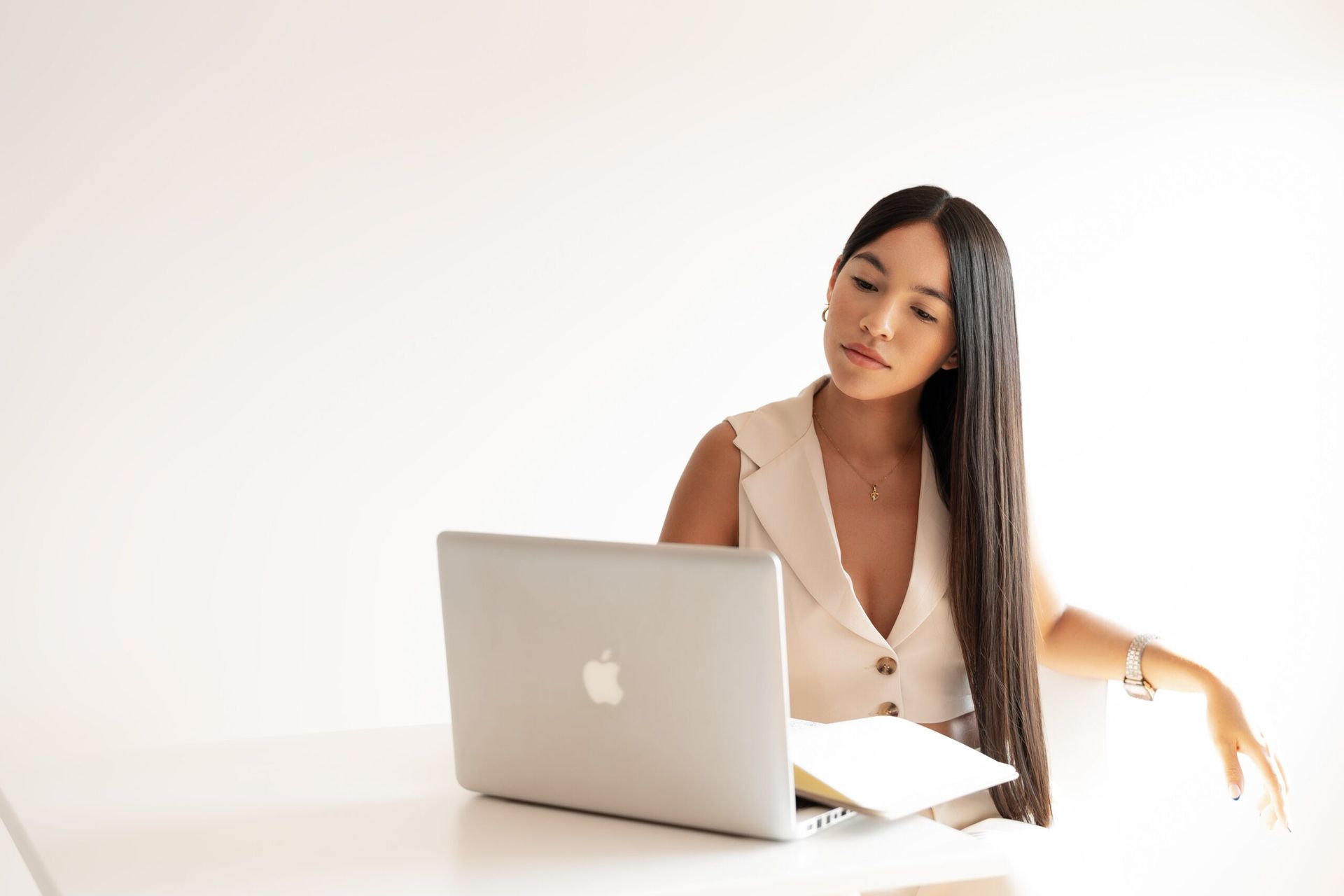 Woman with long dark hair, working on a laptop at a white desk, looking down pensively.