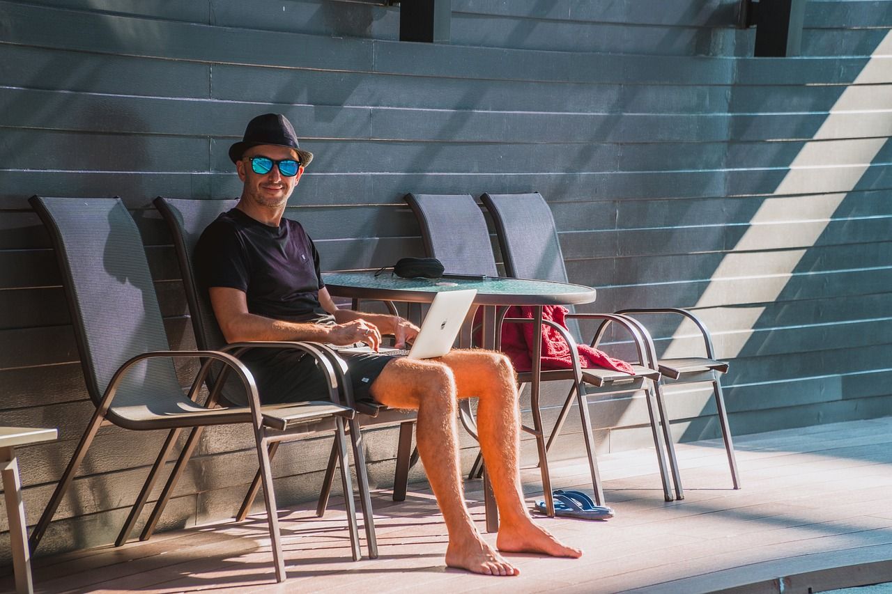 Man in sunglasses and hat working on laptop outside.