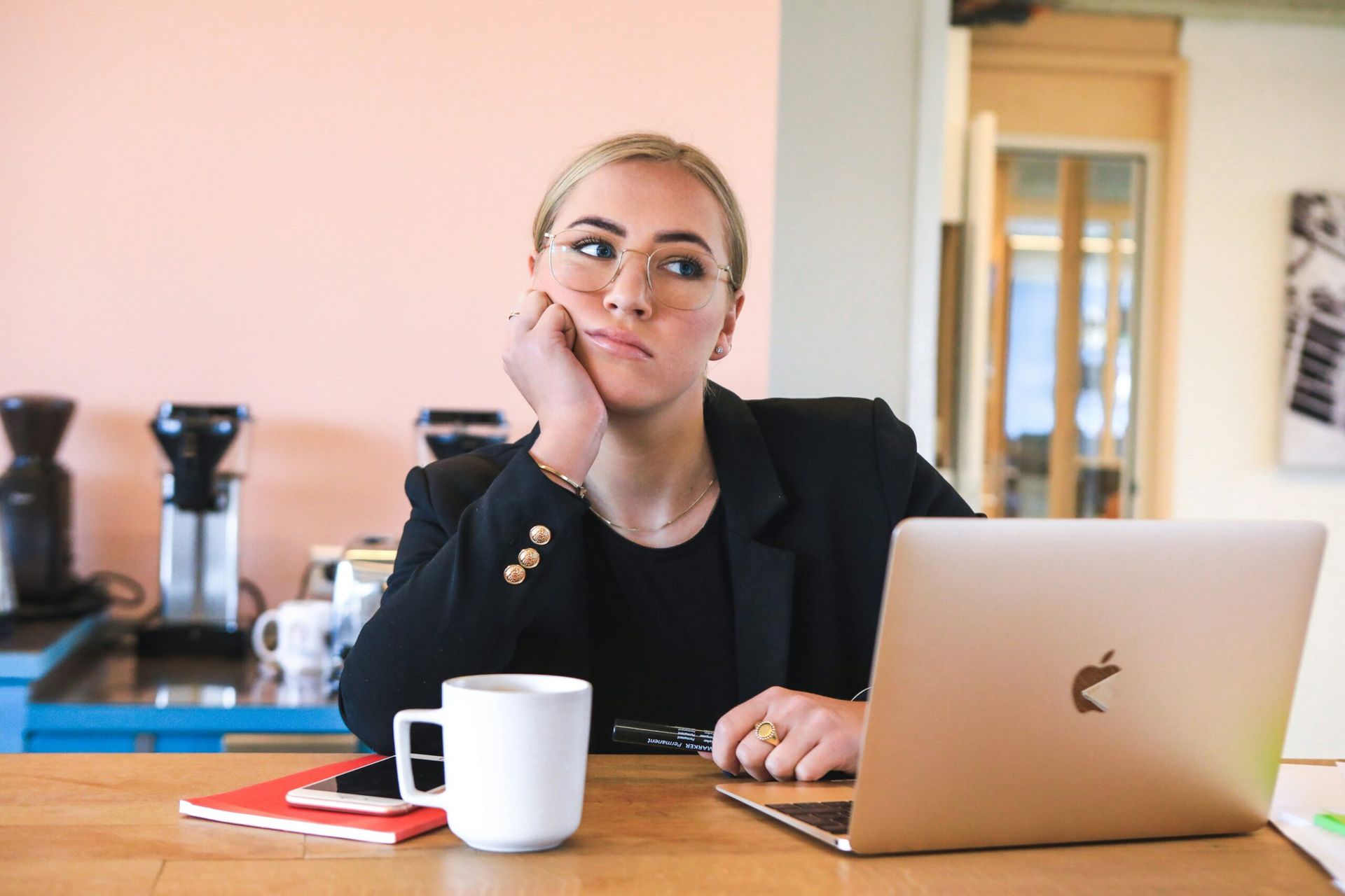 Woman in blazer, bored at desk with laptop, coffee mug, and phone; pink wall background.
