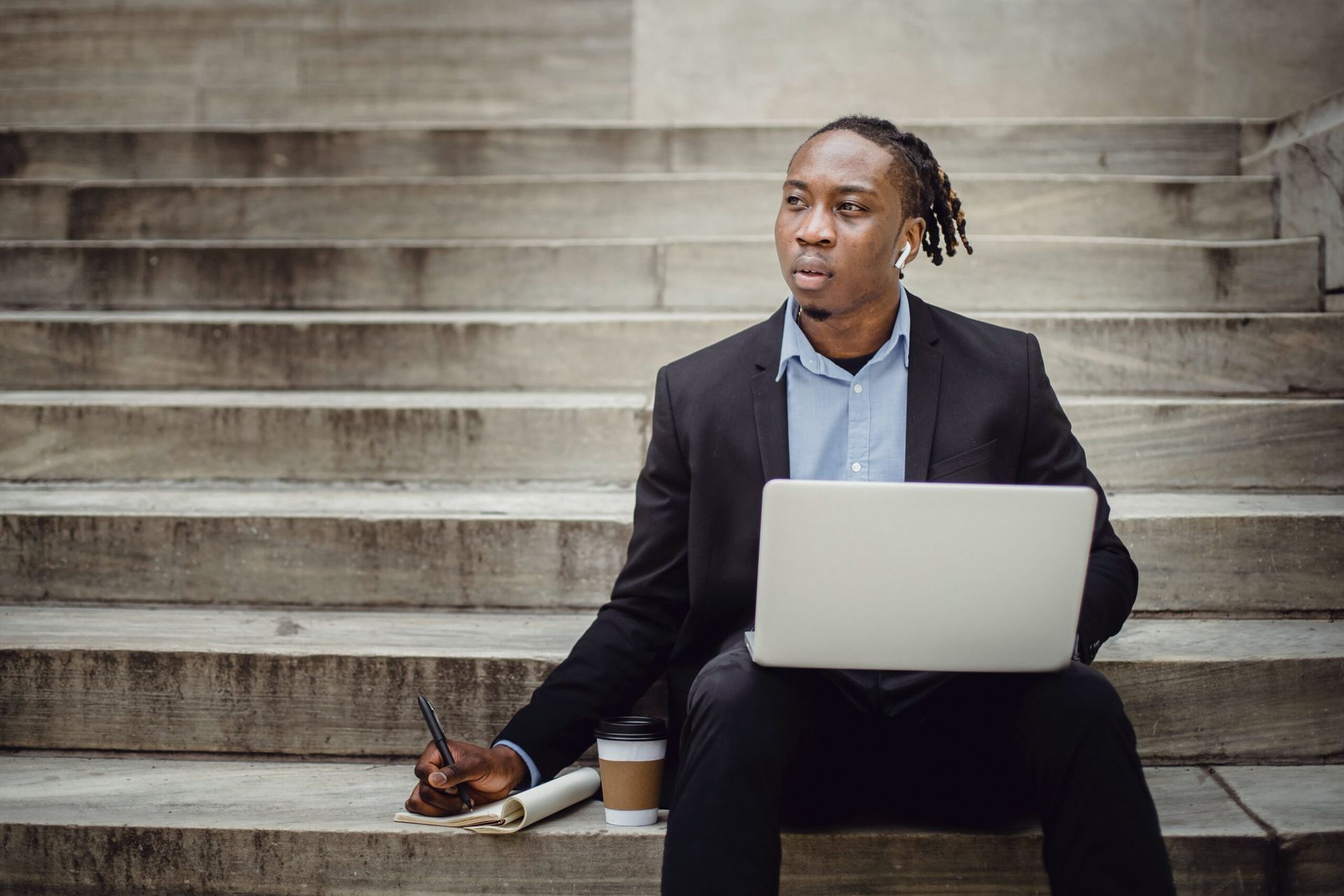 Man in a suit sits on steps, working on a laptop, with a notepad and coffee, looking thoughtful.