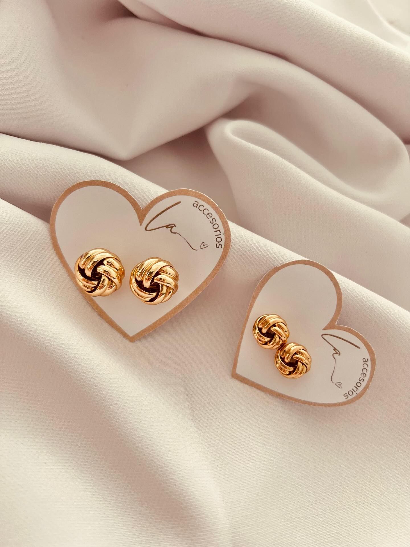 Pendientes de nudo dorado sobre tarjetas con forma de corazón, apoyados sobre tela blanca.