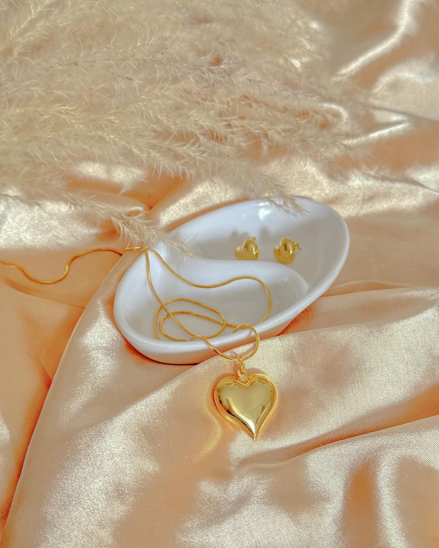 Collar con colgante de corazón dorado y pendientes a juego en plato blanco sobre satén beige.