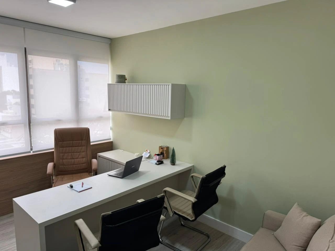 Escritório com mesa, cadeiras e persianas. Paredes verde-claras, mesa branca e móveis bege.