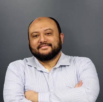 Man with a beard, arms crossed, wearing a striped shirt, against a gray background. Rhames Nordien