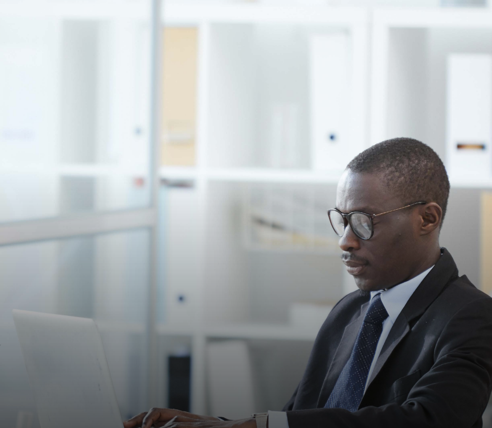 Two people in navy suits, standing in a well-lit office setting. The woman in the front is smiling, hands clasped.